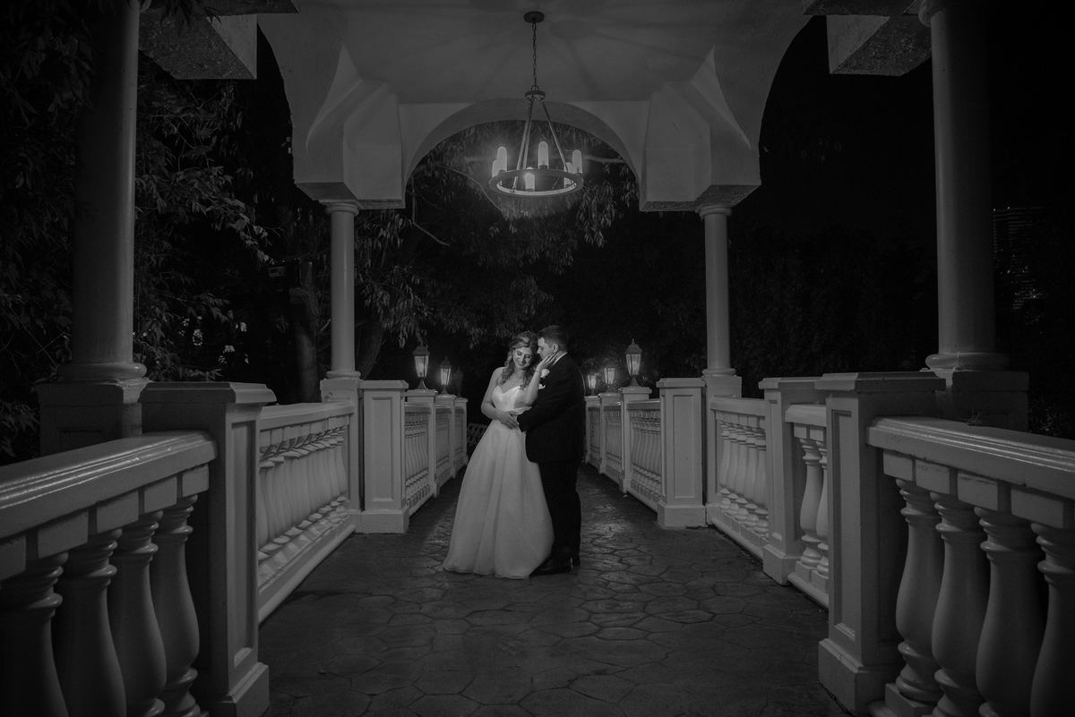 A bride and groom embrace at night beneath a gazebo lit by a chandelier and lanterns, on a stone path between columns.