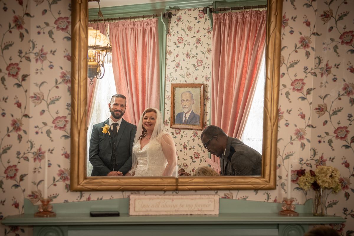 A smiling bride and groom stand in front of an ornate mirror surrounded by floral wallpaper and pink drapes.