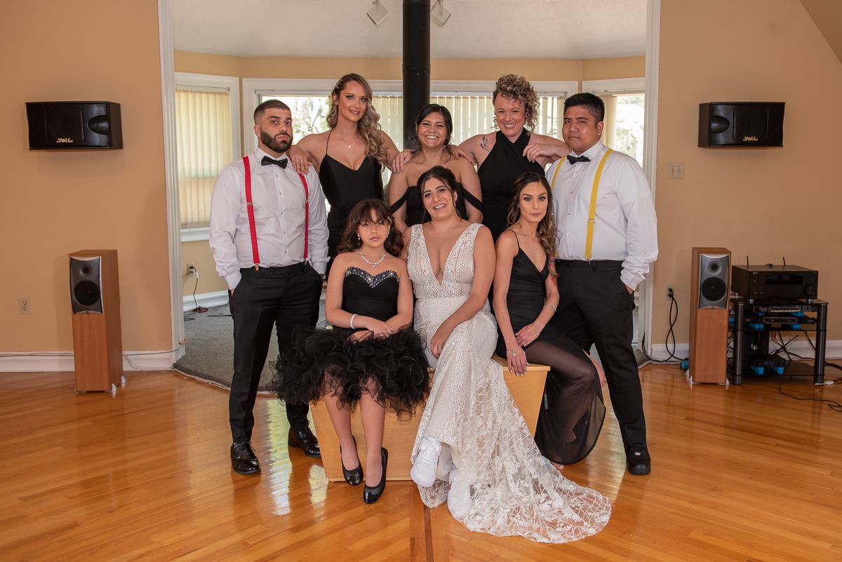 A bride in white is seated with a group of bridesmaids and groomsmen, all posing indoors under natural light.