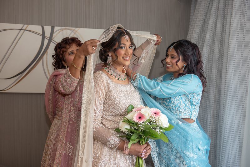 A bride in a cream lehenga is assisted by two women as they drape her with a veil, holding pink and white flowers.