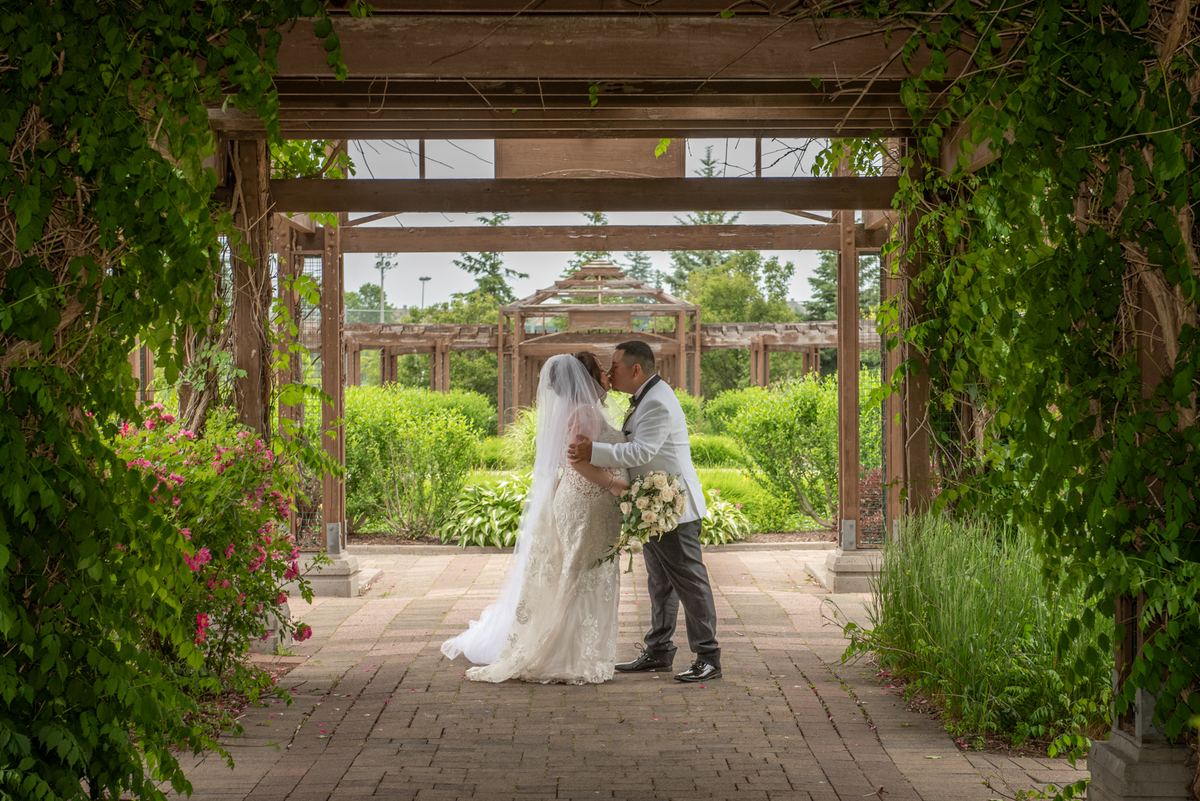 Bride and groom kiss beneath a vine-covered pergola in a garden filled with greenery and pink blossoms.