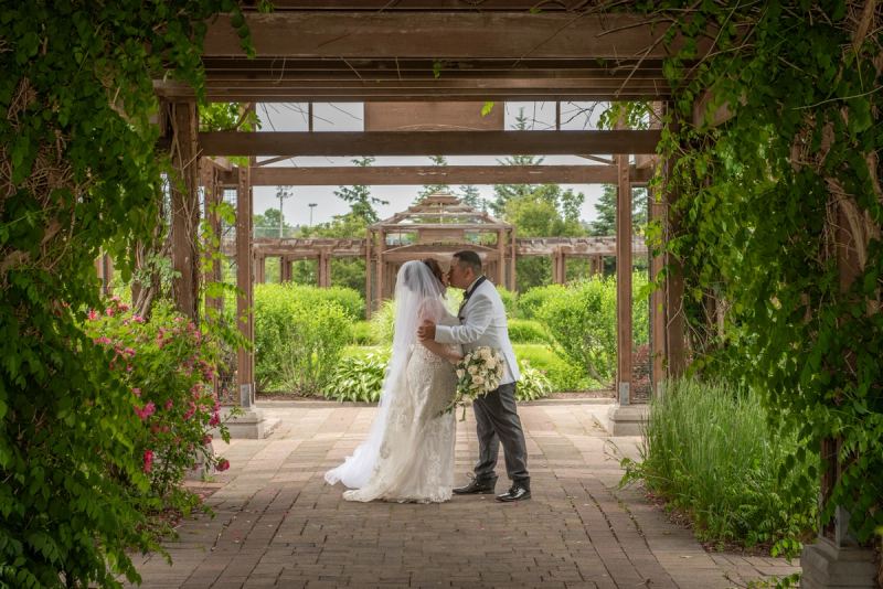 Bride and groom kiss beneath a vine-covered pergola in a garden filled with greenery and pink blossoms.