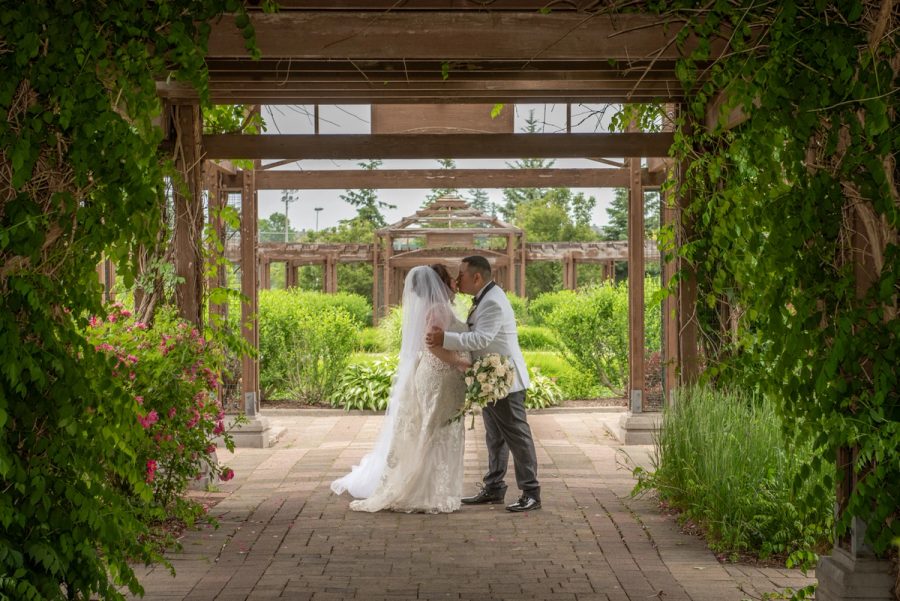 Bride and groom kiss beneath a vine-covered pergola in a garden filled with greenery and pink blossoms.