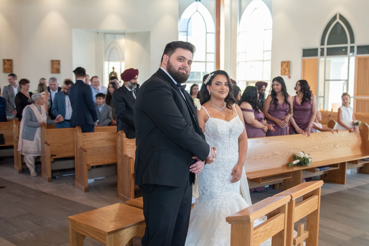 A bride and groom smile at the altar as guests in wooden pews watch during a ceremony illuminated by natural light.