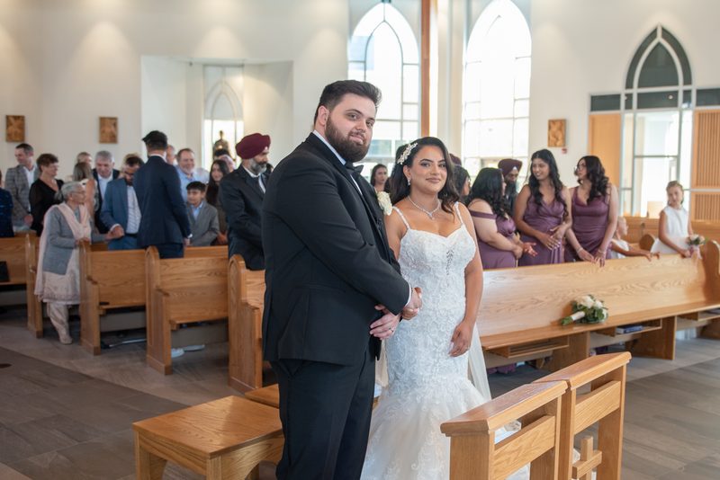 A bride and groom smile at the altar as guests in wooden pews watch during a ceremony illuminated by natural light.