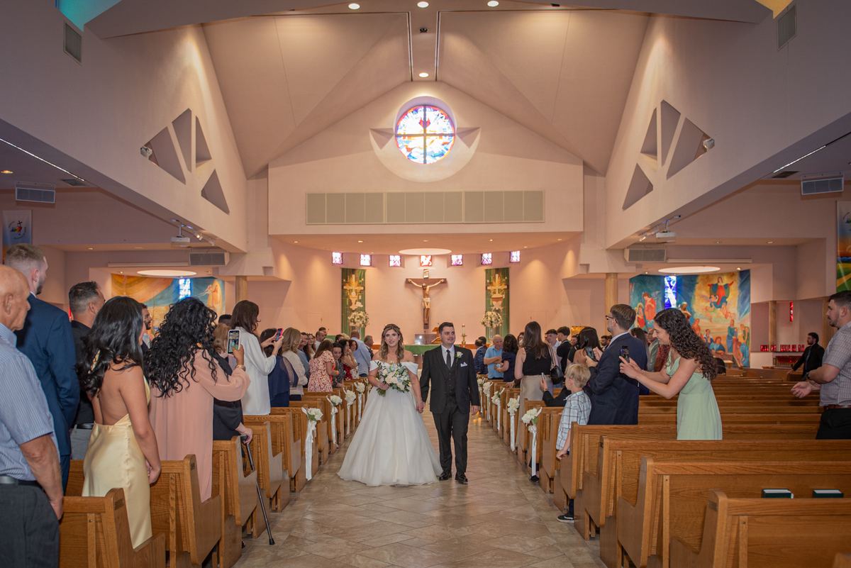 A bride and groom walk down the aisle of a modern church toward the altar, surrounded by guests in wooden pews.