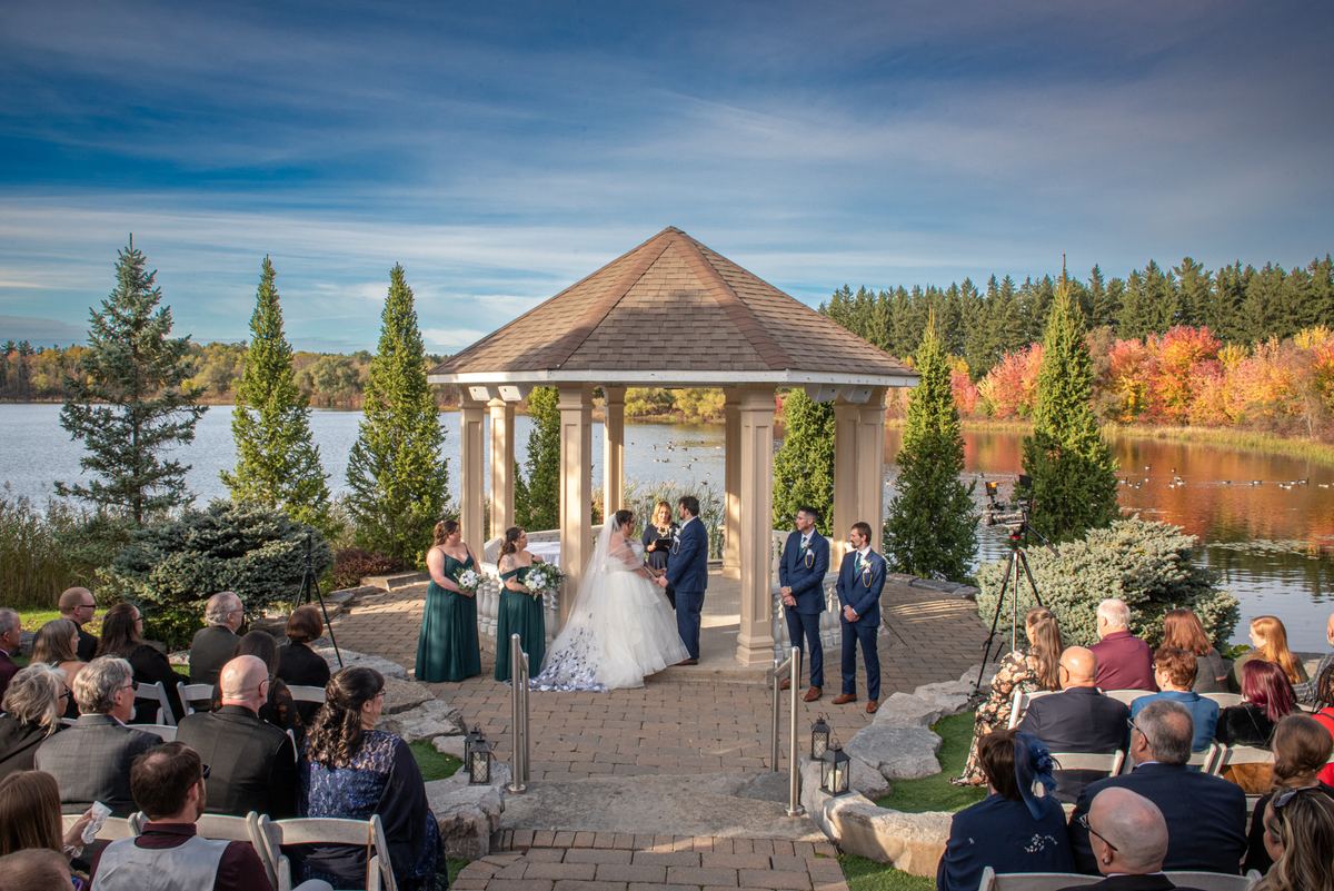 A bride and groom stand in a gazebo by a lake surrounded by guests as autumn leaves frame the ceremony.