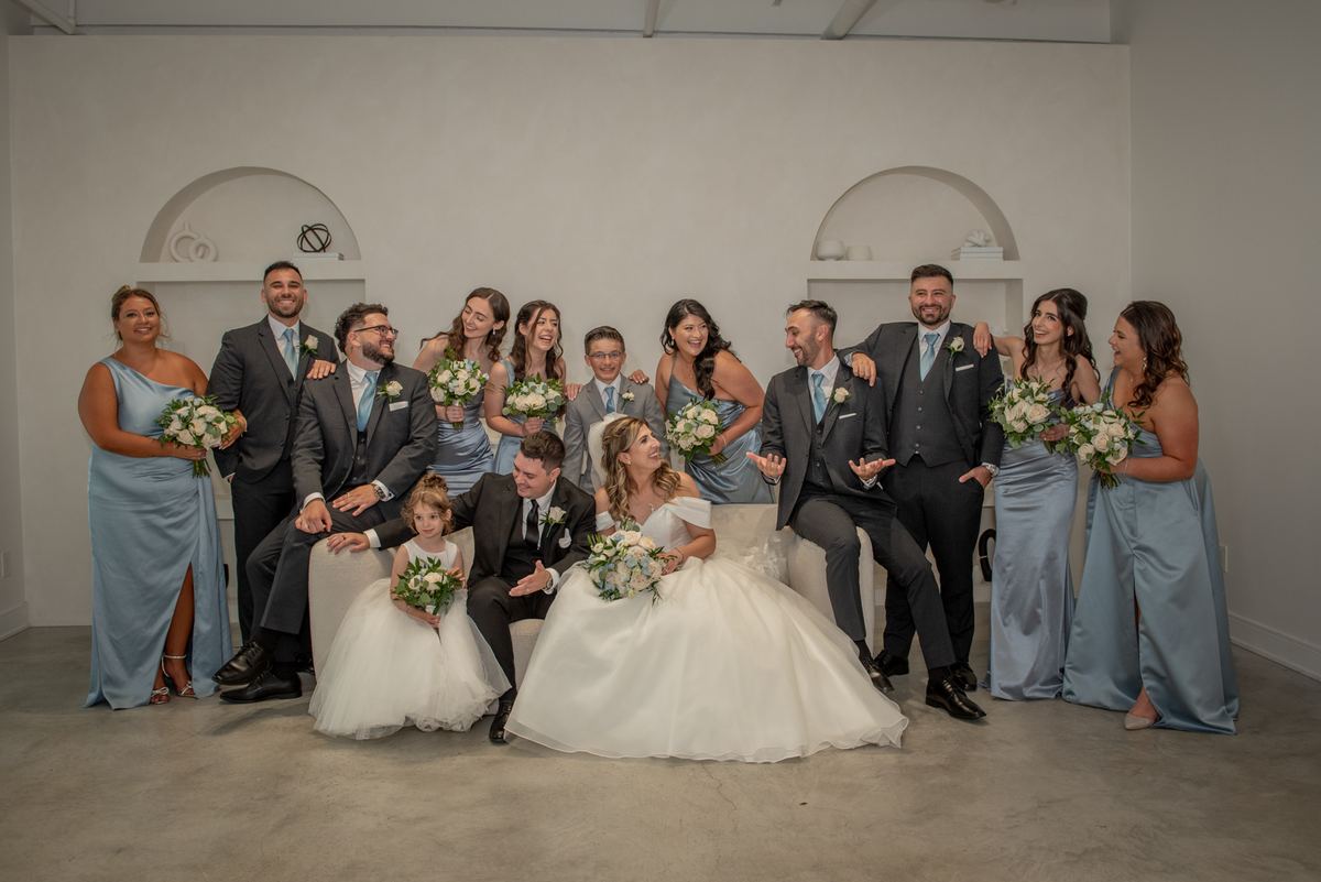 A joyful wedding party poses together in a bright space, the bride and groom front and center with smiling bridesmaids.