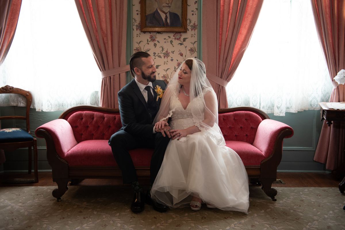 A newlywed couple in formal attire sit closely on a red sofa, gazing at each other in an ornate room with floral wallpaper.