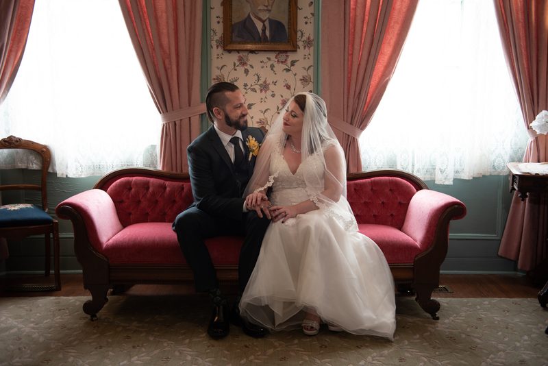 A newlywed couple in formal attire sit closely on a red sofa, gazing at each other in an ornate room with floral wallpaper.
