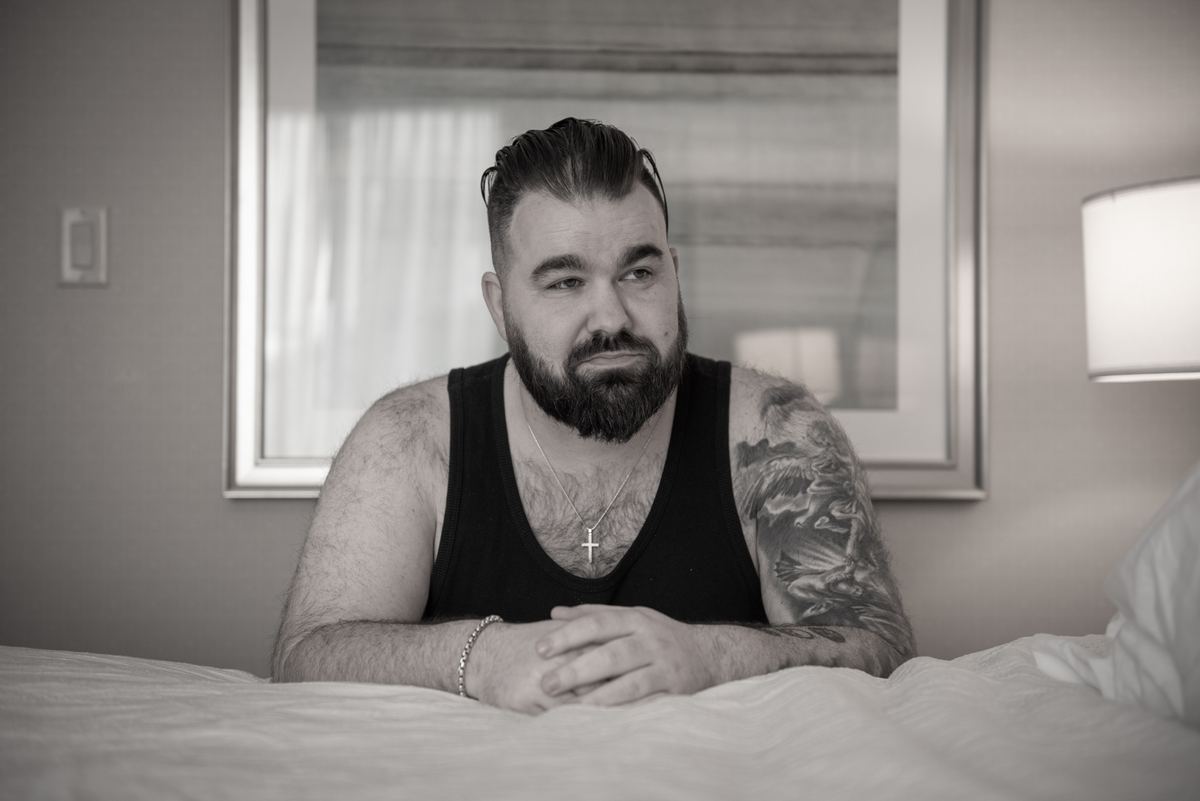 A bearded man with tattoos sits pensively on a bed in soft lighting, hands clasped and gazing off-camera.