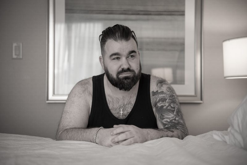 A bearded man with tattoos sits pensively on a bed in soft lighting, hands clasped and gazing off-camera.