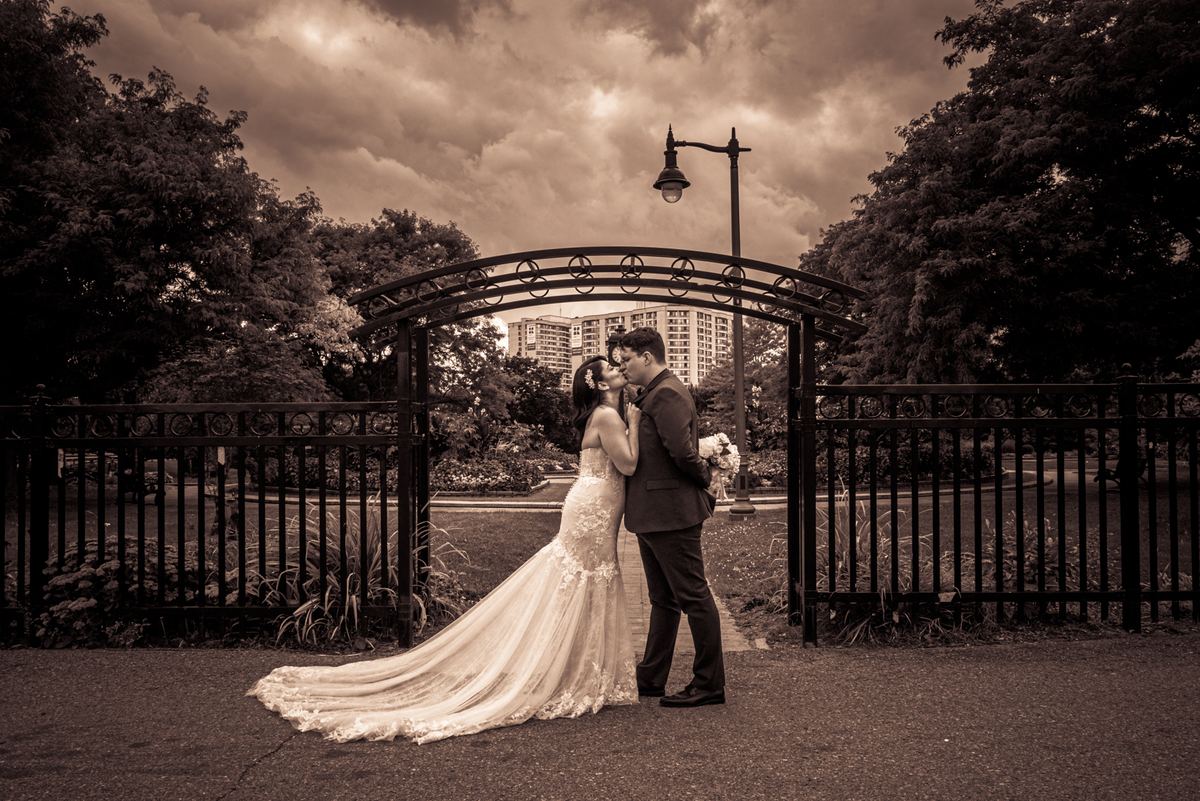 A newlywed couple kisses beneath an ornate metal archway in a park, surrounded by trees and a dramatic sky.