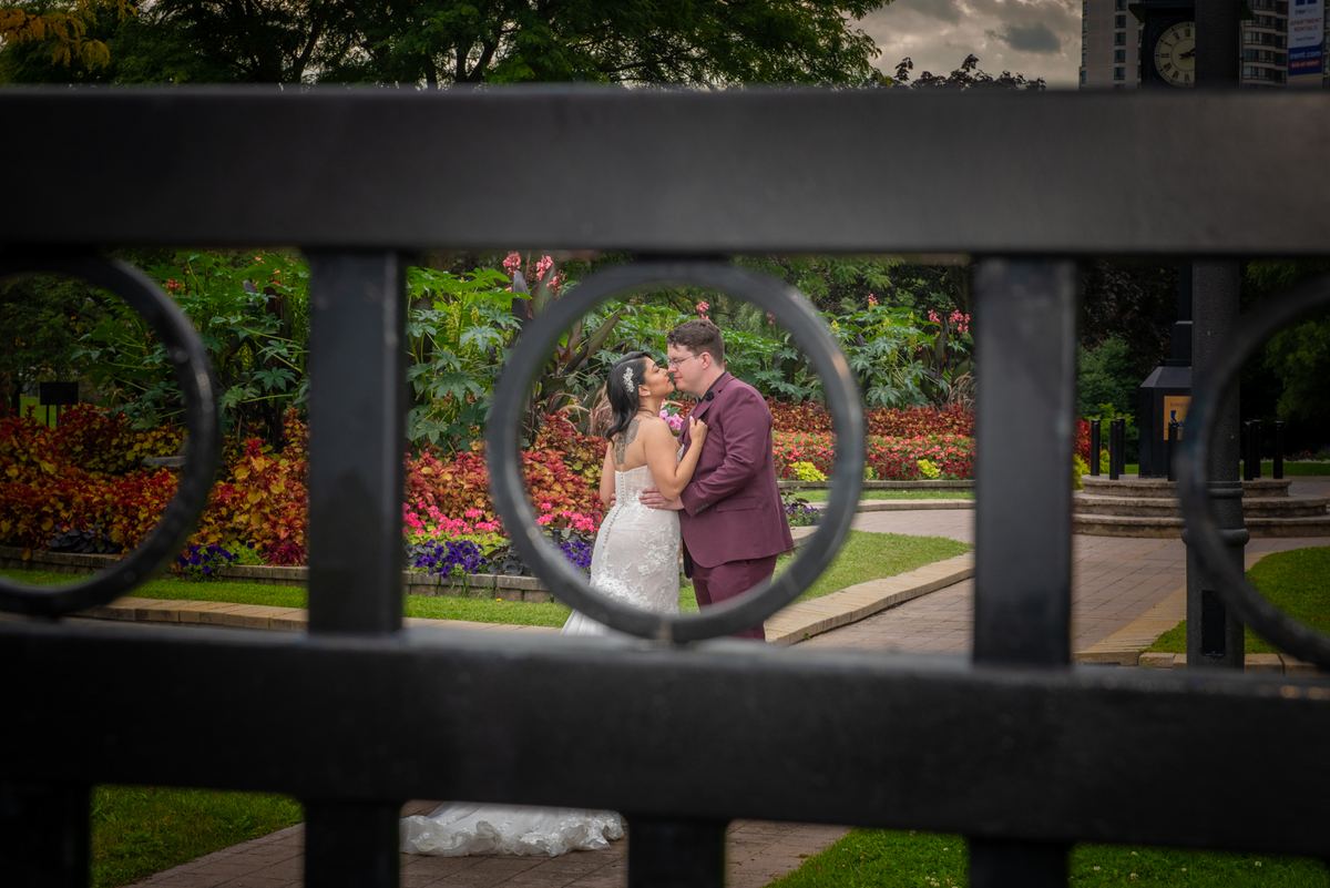 A bride and groom kiss in a garden surrounded by black iron fence circles, with autumn leaves and flowers in the background.