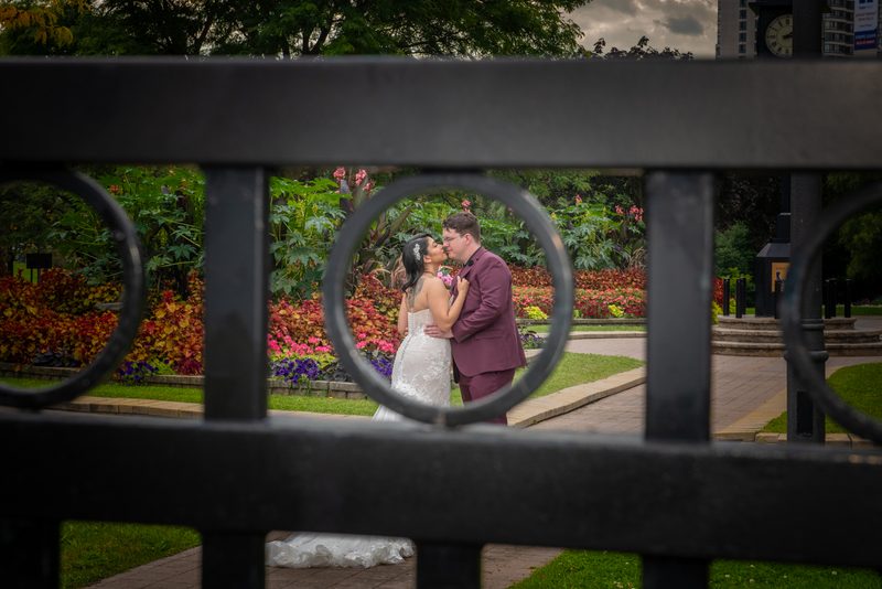 A bride and groom kiss in a garden surrounded by black iron fence circles, with autumn leaves and flowers in the background.