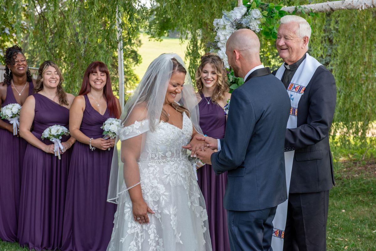 A bride and groom exchange vows under a willow tree with their bridal party and officiant in warm daylight.