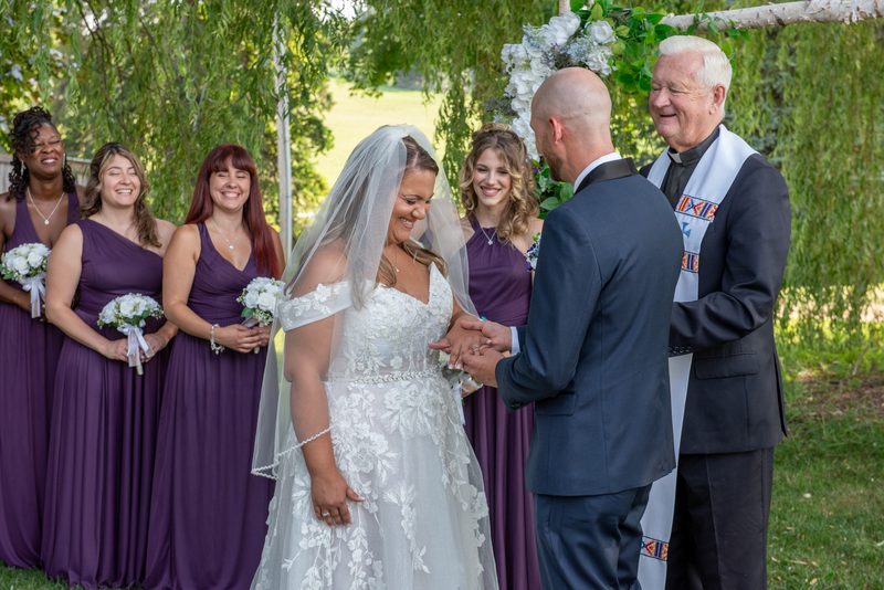 A bride and groom exchange vows under a willow tree with their bridal party and officiant in warm daylight.
