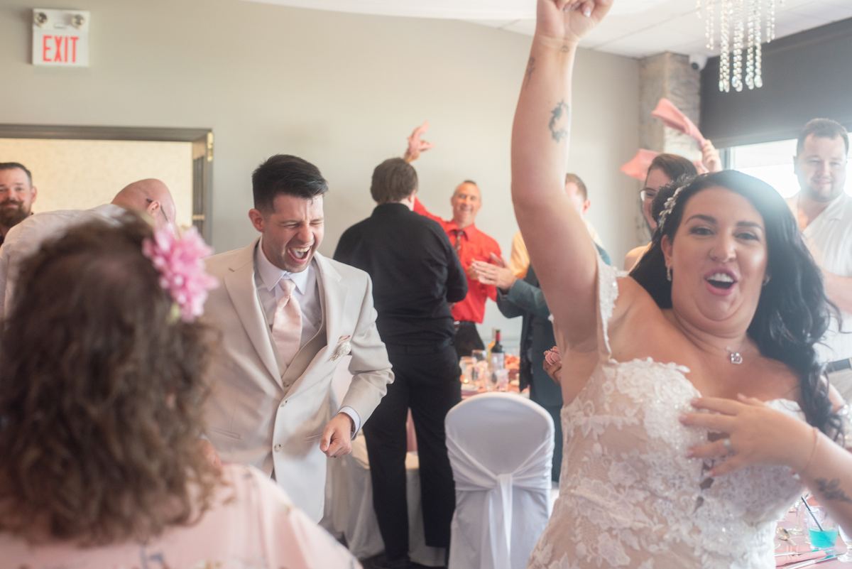 Bride in lace dress dances exuberantly with raised arm, groom laughs beside her at lively reception.