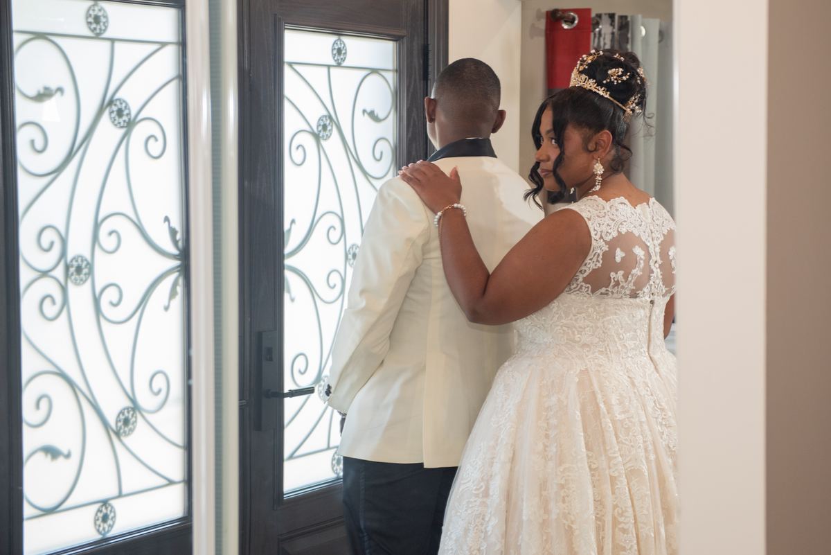Bride in lace gown and tiara stands back-to-back with groom in white jacket near ornate glass door, hands touching.