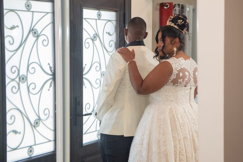 Bride in lace gown and tiara stands back-to-back with groom in white jacket near ornate glass door, hands touching.