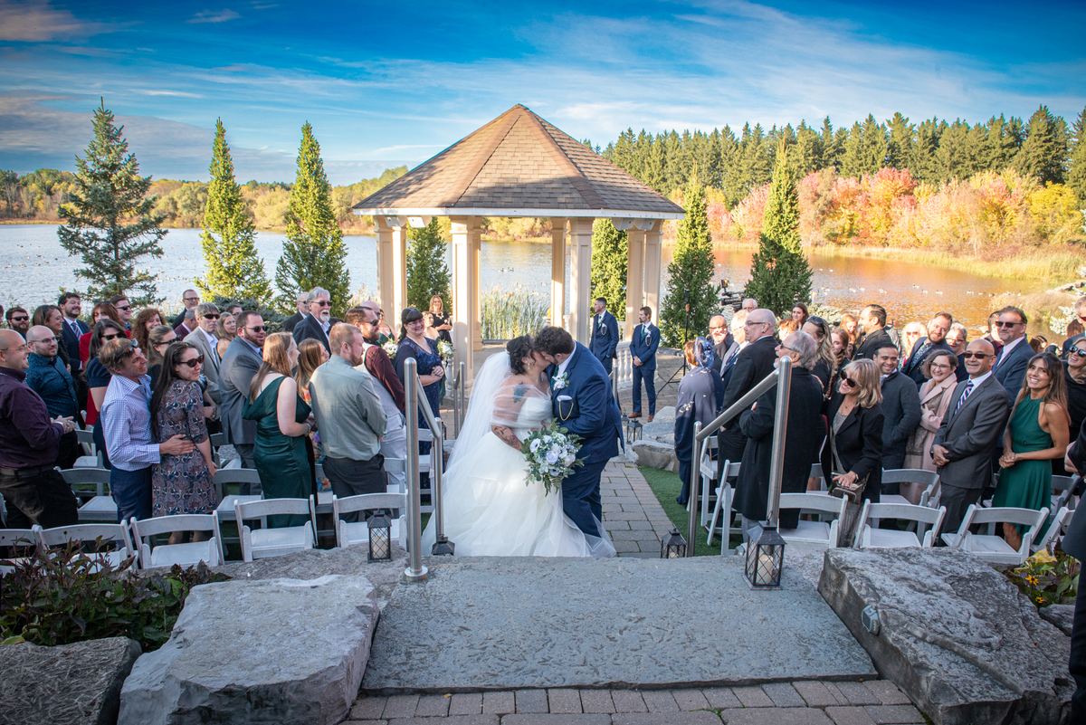 A bride and groom kiss at an outdoor altar surrounded by guests in white chairs, with a lake, autumn trees.