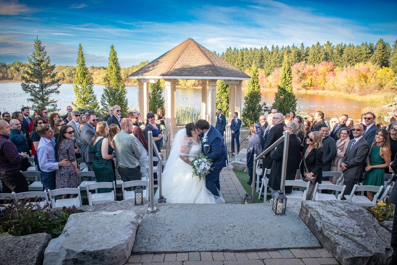 A bride and groom kiss at an outdoor altar surrounded by guests in white chairs, with a lake, autumn trees.