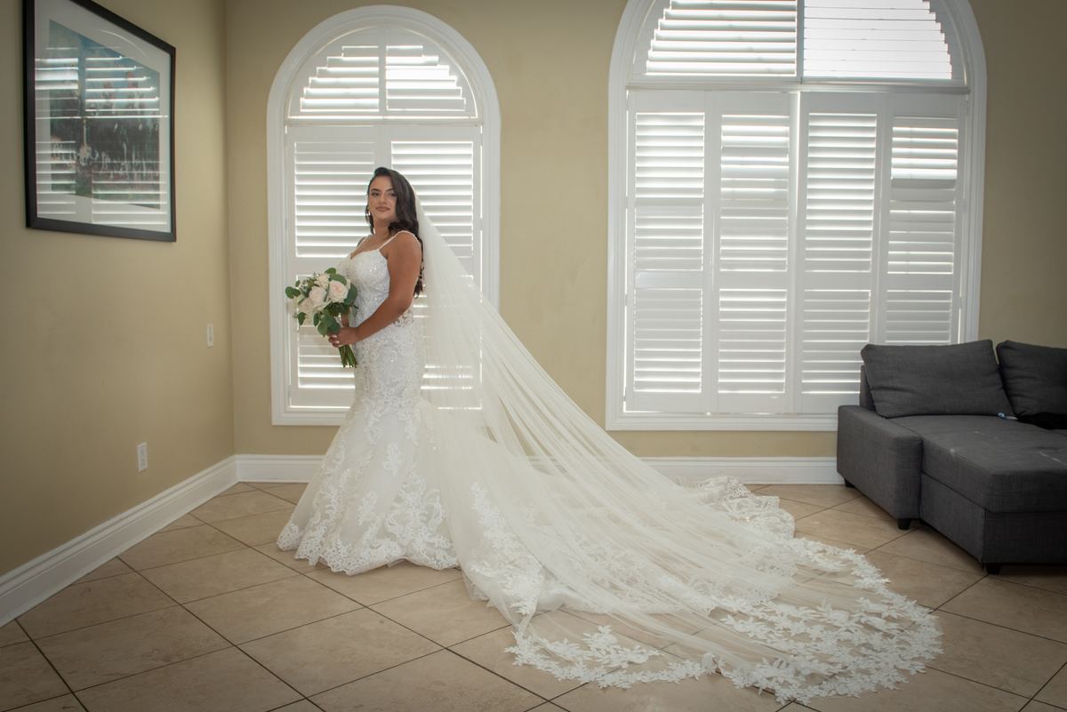 A bride in an elaborate lace gown and veil stands near arched windows, holding a bouquet of white and green flowers.