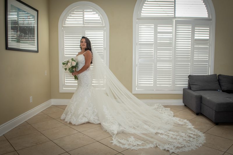 A bride in an elaborate lace gown and veil stands near arched windows, holding a bouquet of white and green flowers.
