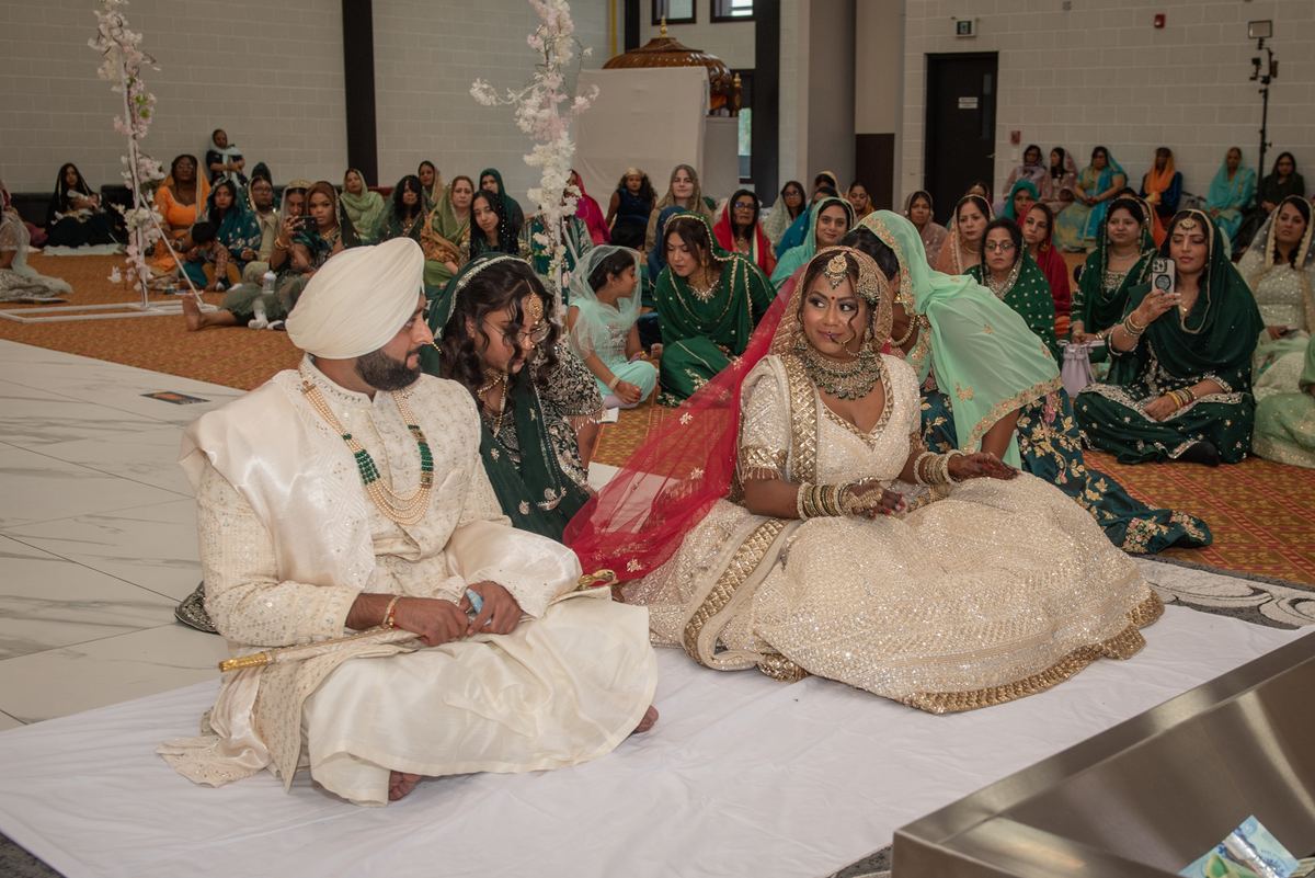 Bride in ornate lehenga and groom in sherwani sit calmly during ceremony surrounded by guests in traditional attire.