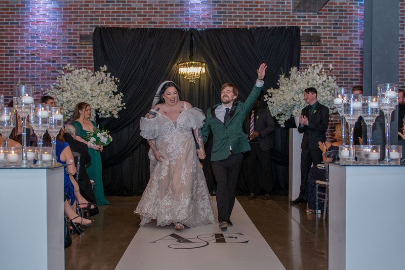 A groom raises his hand in celebration as he and his bride walk down a candlelit aisle between rows of applauding guests.