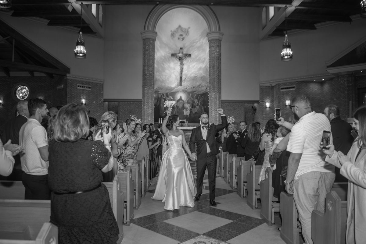 Bride and groom walk down the aisle with arms raised as guests cheer on their phones during a church wedding.