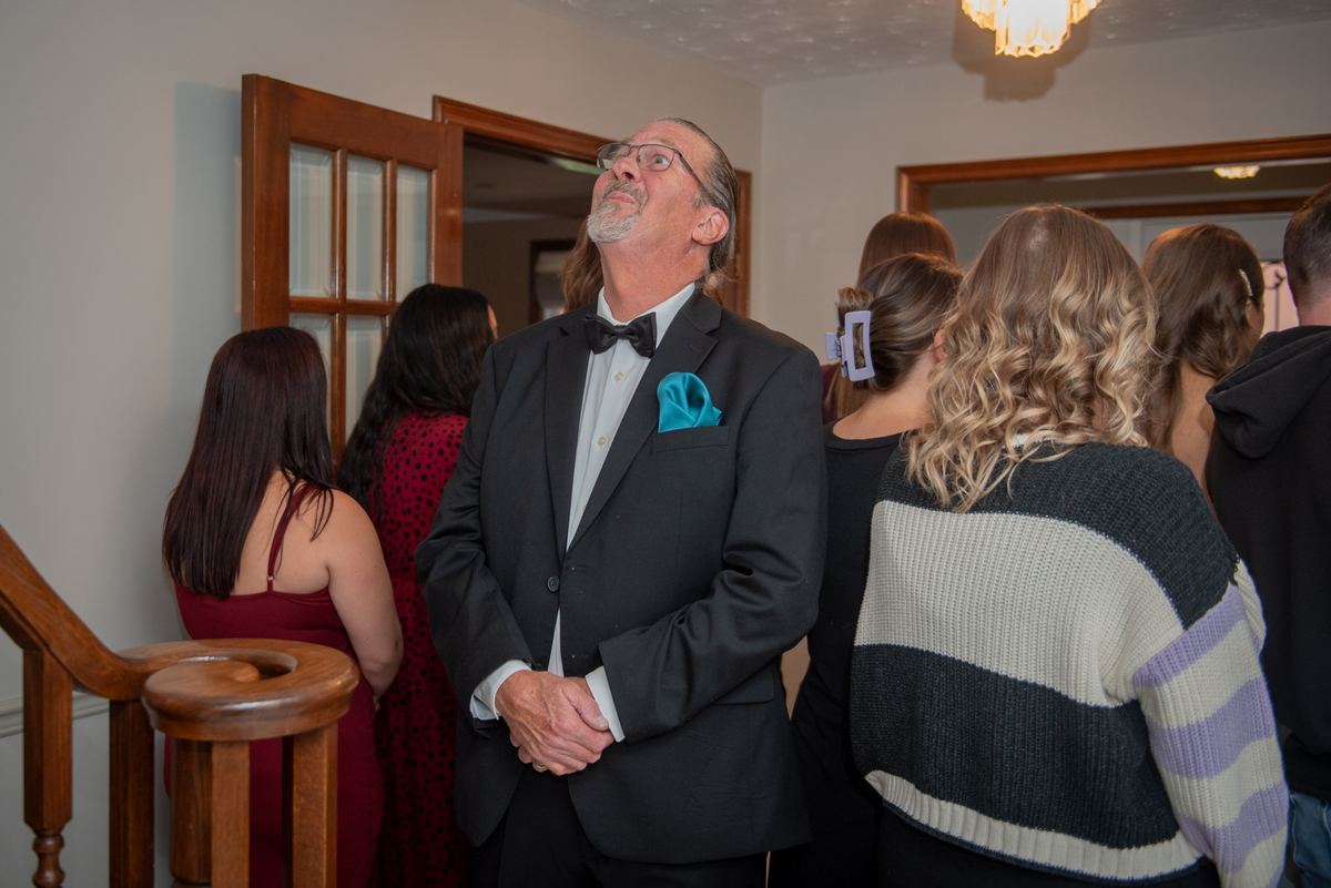 A man in a tuxedo stands with guests below a chandeliered staircase, all looking upward in anticipation.