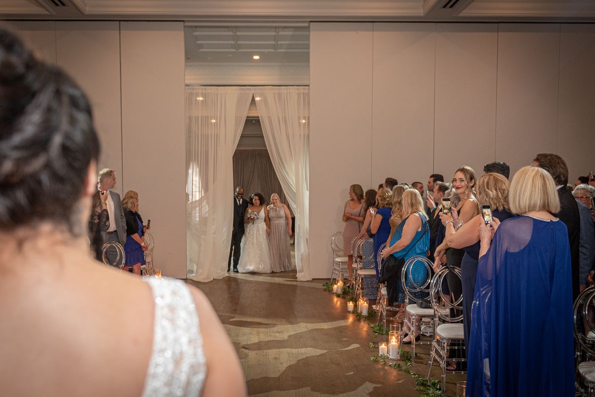 A bride and her attendants walk down a candlelit aisle toward the ceremony space, framed by white curtains.