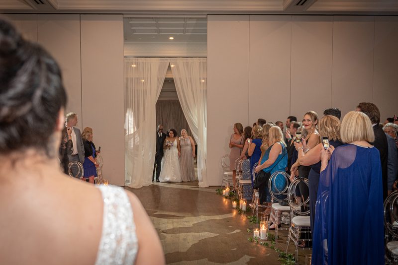 A bride and her attendants walk down a candlelit aisle toward the ceremony space, framed by white curtains.