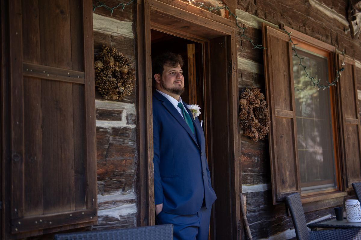 A groom in navy suit and green tie stands expectantly at a rustic cabin door, illuminated by soft natural light.