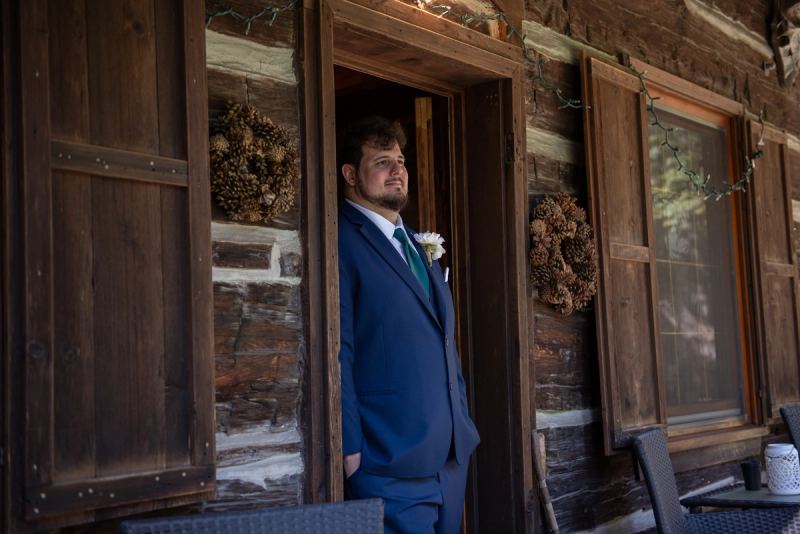 A groom in navy suit and green tie stands expectantly at a rustic cabin door, illuminated by soft natural light.