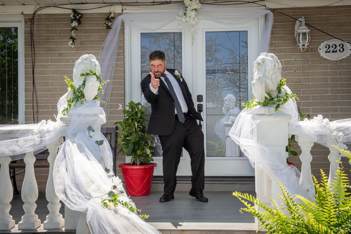 A groom in a dark suit points at the camera with a grin, framed by veiled statues and greenery on a porch.