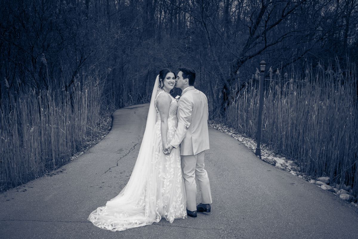 A bride and groom stand on a winding path, the groom kissing her cheek as she smiles warmly in late afternoon light.