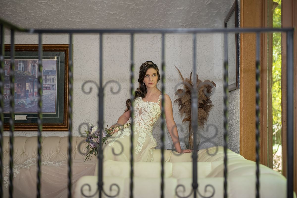 A bride in a lace gown stands by an iron railing, holding flowers near a window-lit room with furniture and art.