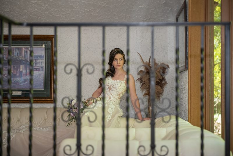 A bride in a lace gown stands by an iron railing, holding flowers near a window-lit room with furniture and art.