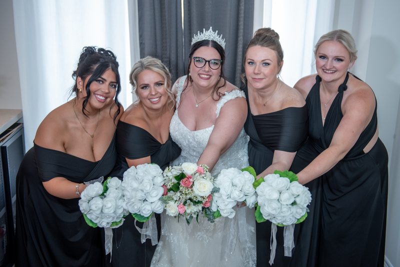 A bride in a white gown and tiara poses with four bridesmaids in black dresses, holding rose bouquets near sheer curtains.