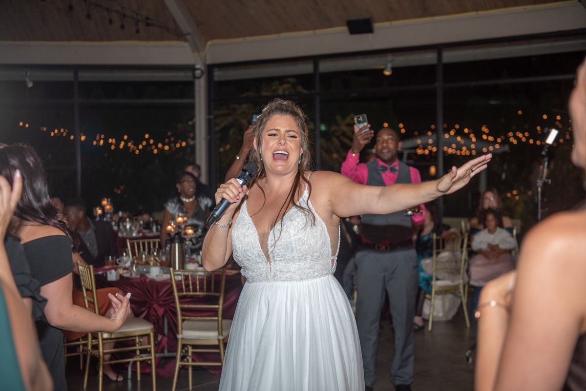 A bride in a white gown sings passionately at a nighttime reception with guests clapping and recording.