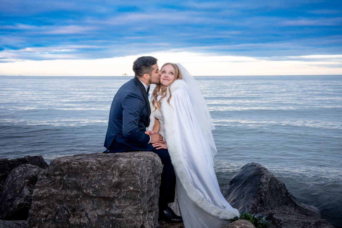 A bride and groom seated on rocks by the sea, sharing a kiss in soft, blue-toned light reflecting off calm water.
