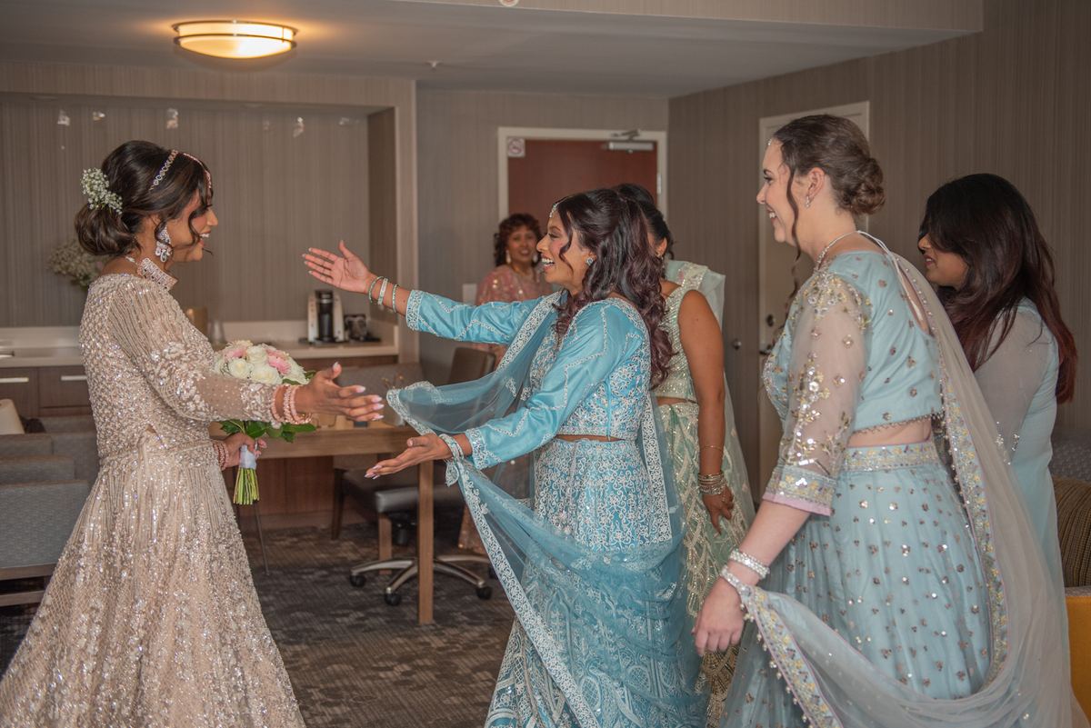 A bride in a beige lehenga smiles and gestures toward laughing bridesmaids in light blue, holding a bouquet.