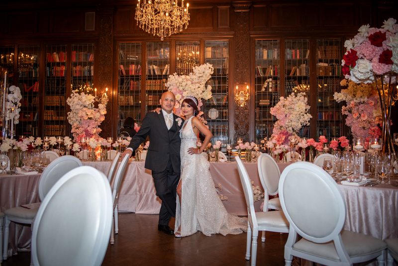 A bride and groom smile in a grand banquet hall adorned with flowers and candles, surrounded by guests.
