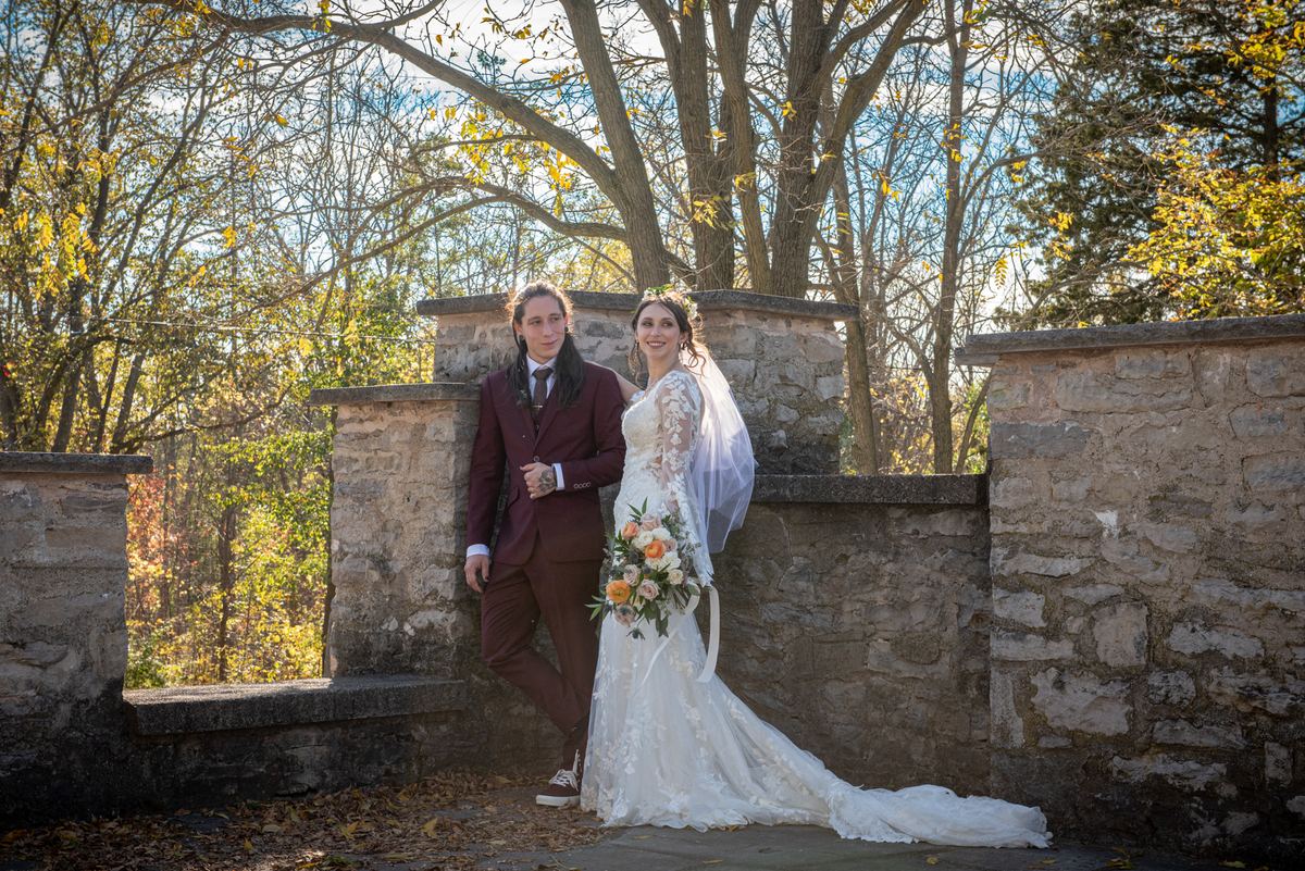 A bride in a lace gown and veil with her groom in a burgundy suit, posing joyfully against a rustic stone wall.