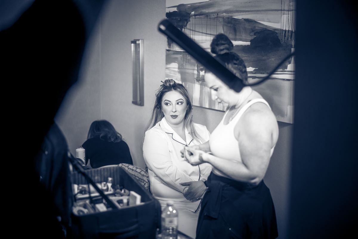 A woman in a white robe is being assisted by another with her hair and accessories, curling iron visible.