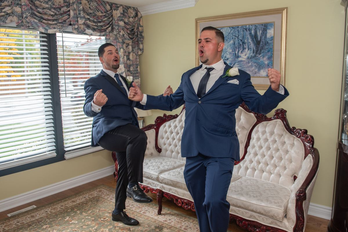 Two men in navy suits dance joyfully in a warmly lit room with ornate curtains and a tufted sofa visible.