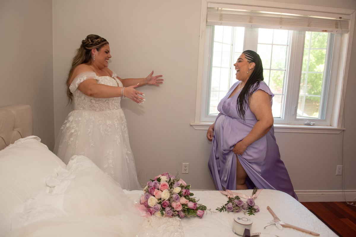 Two brides laughing in a bedroom, one lifting her dress to show her pregnant belly, with natural light.
