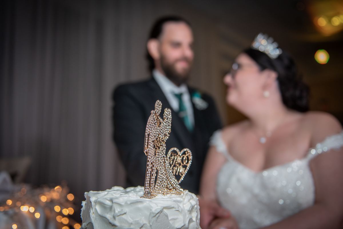 Bride and groom stand behind wedding cake with sparkling topper, gazing at each other in warm lighting.